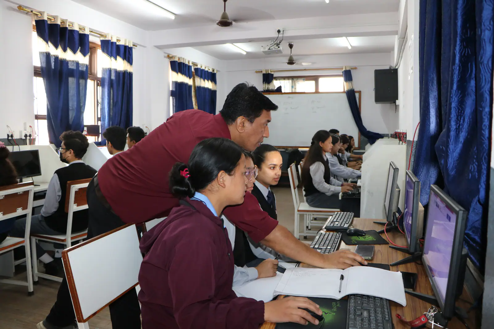 Students at rows of desktop machines in the computer laboratory, a teacher leaning over one screen