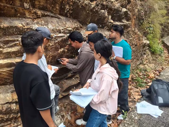 Students taking notes at an exposed rock face, one holding a clipboard, packs on the ground