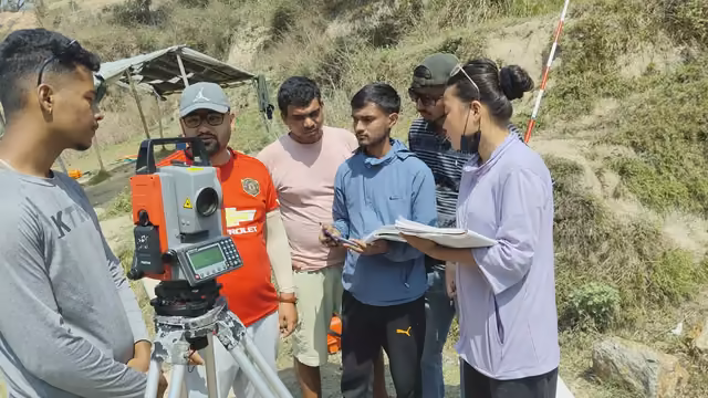 Students gathered round a total station on a hillside at survey camp, a tarpaulin shelter pitched behind