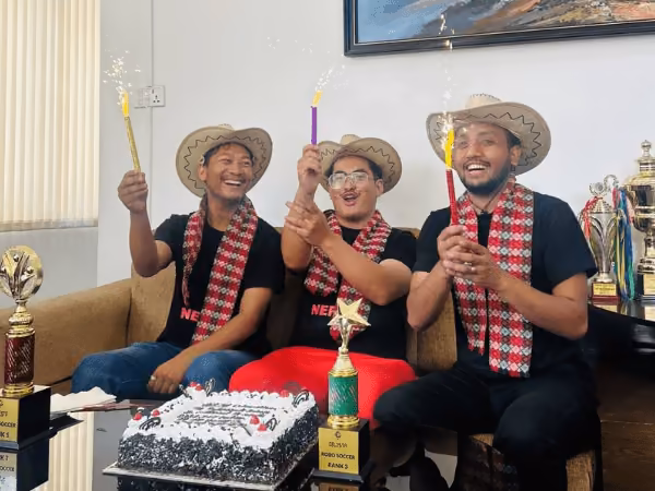 Three students in hats with a cake and trophies after a robot football competition