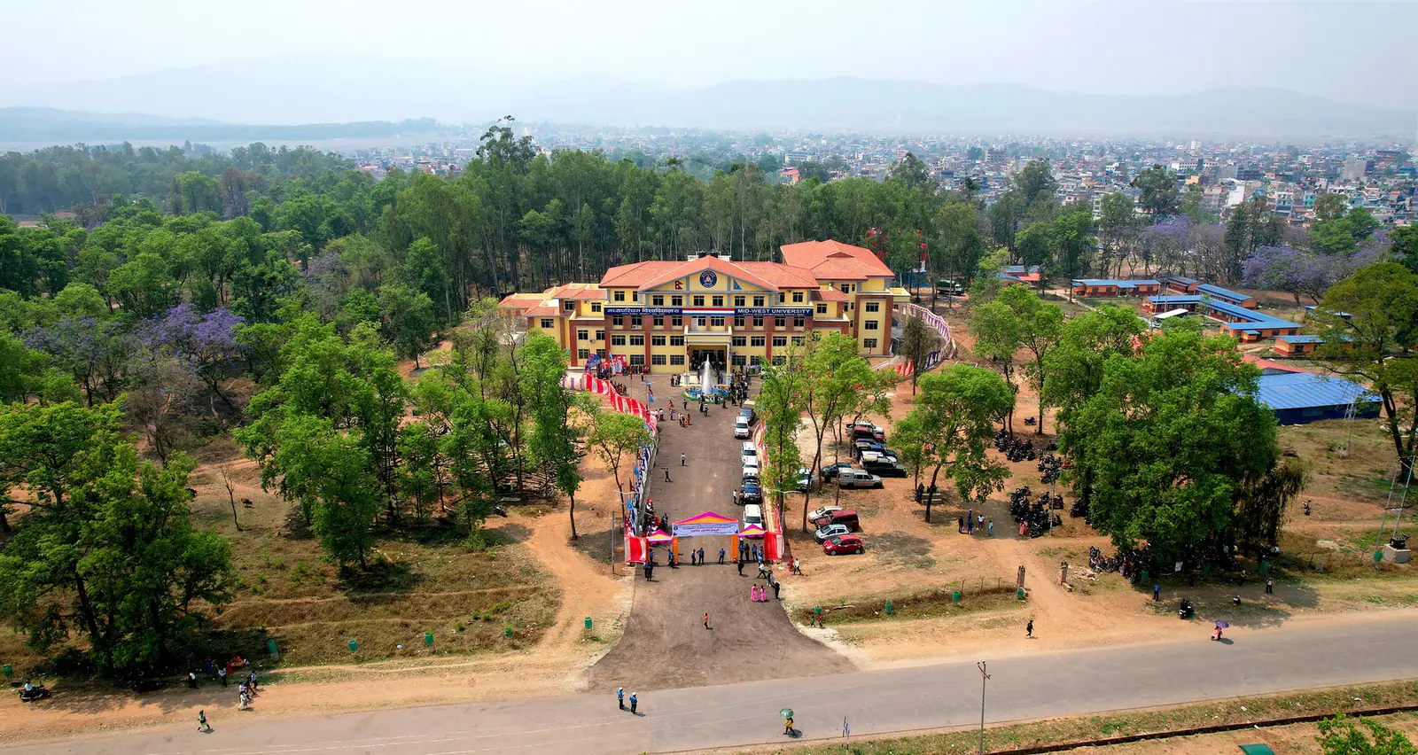 Aerial view of the red-roofed campus blocks among trees, a drive of parked cars leading in