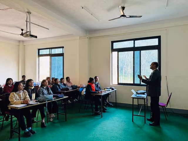 A teacher addressing students at wooden benches in a green-floored classroom with wide windows