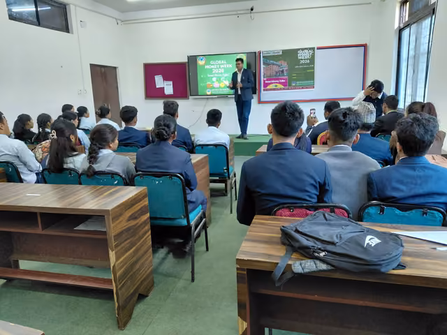 A full classroom watching a projected slide, bags on the benches at the back of the room