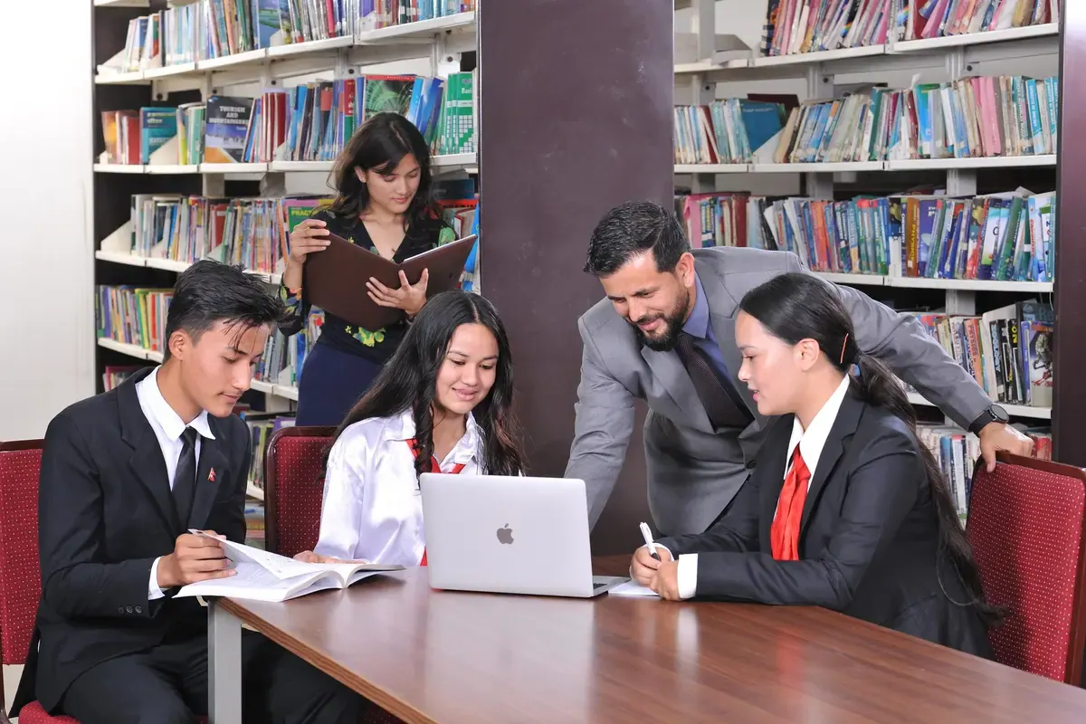 Students working at a laptop and open books at a library table, shelves of books all around