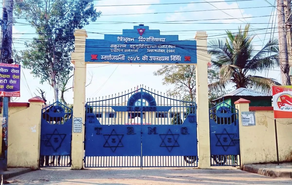 The blue iron campus gate under its Nepali nameboard, palms rising behind the pillars
